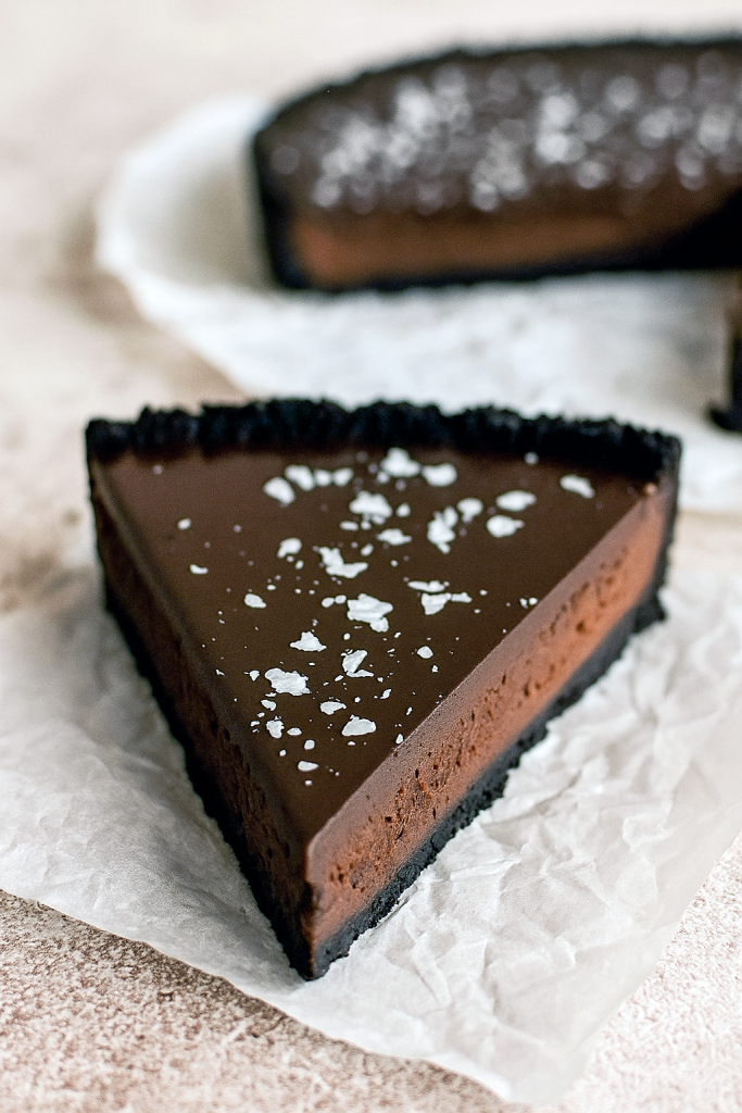 A slice of glossy dark chocolate ganache tart resting on a porcelain plate, its crisp chocolate cookie crust contrasted by a silky, mirror‑like ganache surface, dusted with flaky sea salt.