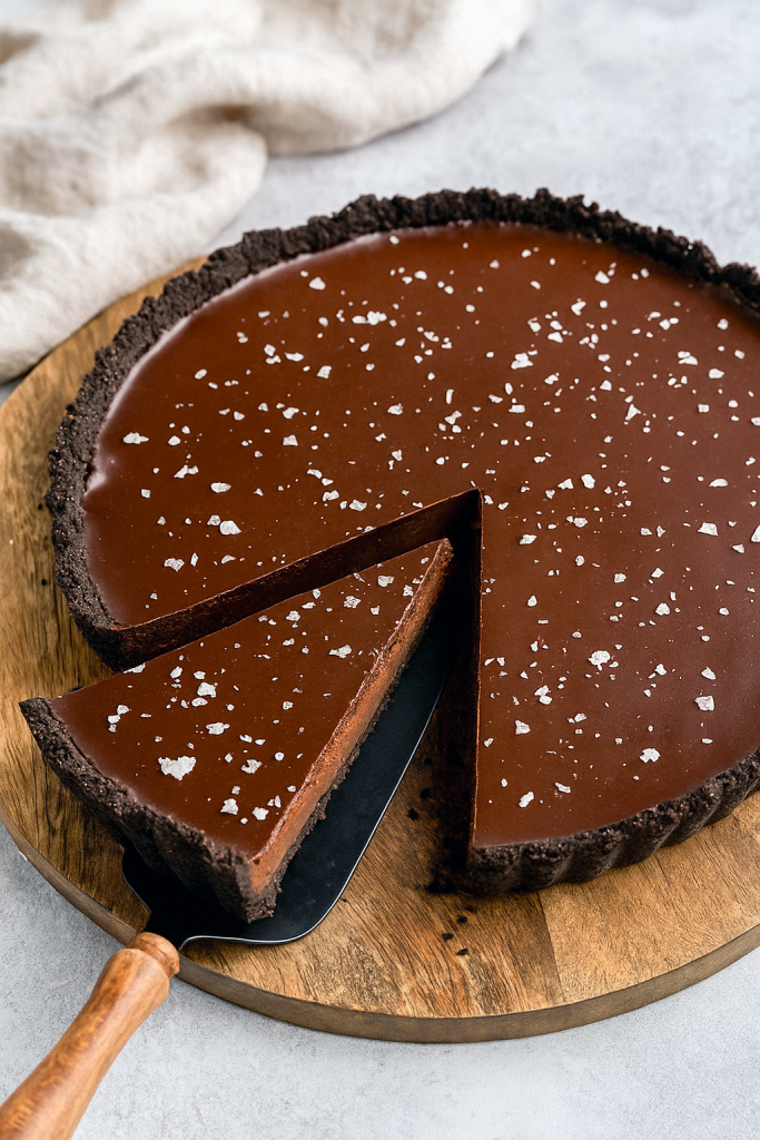 A slice of glossy dark chocolate ganache tart resting on a porcelain plate, its crisp chocolate cookie crust contrasted by a silky, mirror‑like ganache surface, dusted with flaky sea salt.