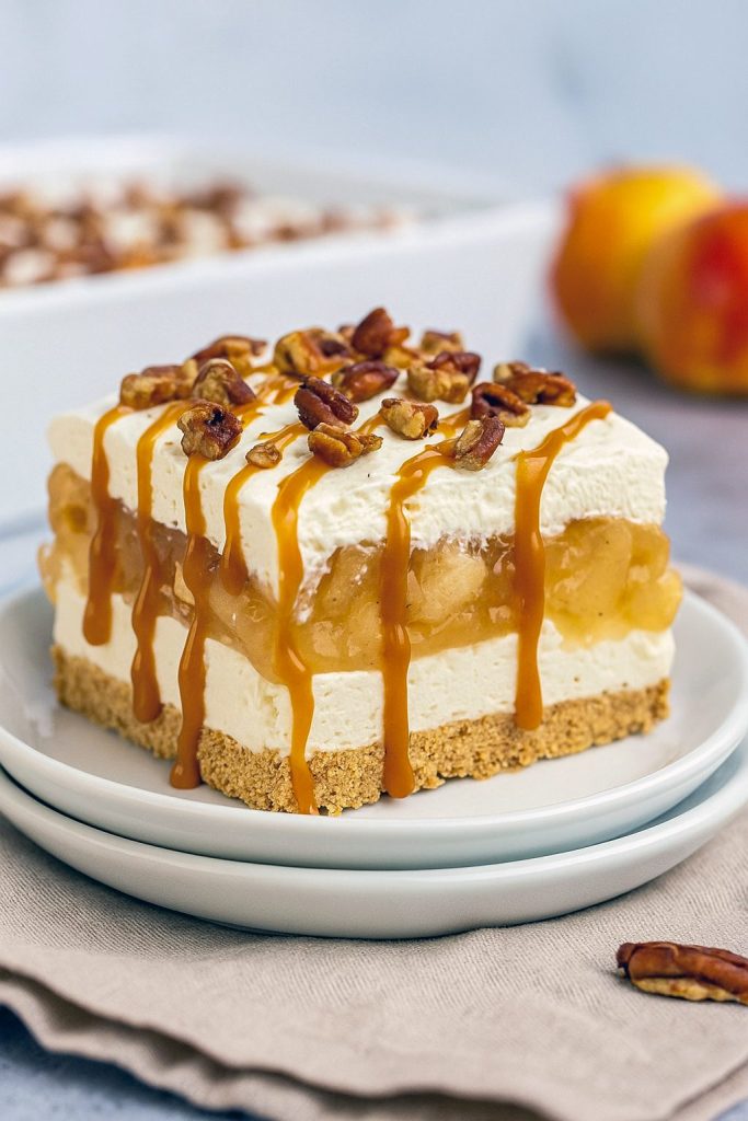 A glass dish filled with layered apple lasagna dessert: a golden graham cracker crust, creamy white cheesecake layer, juicy apple filling, topped with whipped cream and chopped pecans.