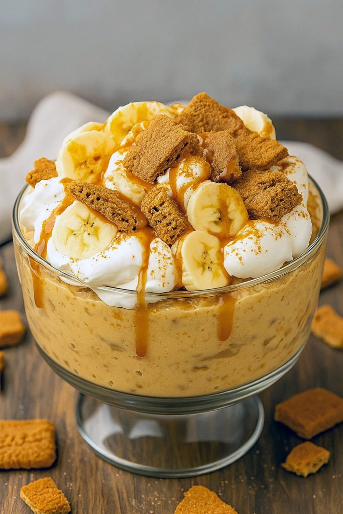 Trifle bowl with alternating layers of creamy vanilla pudding, banana slices, crushed Biscoff cookies and glossy caramel drizzle, topped with cookie crumbs.