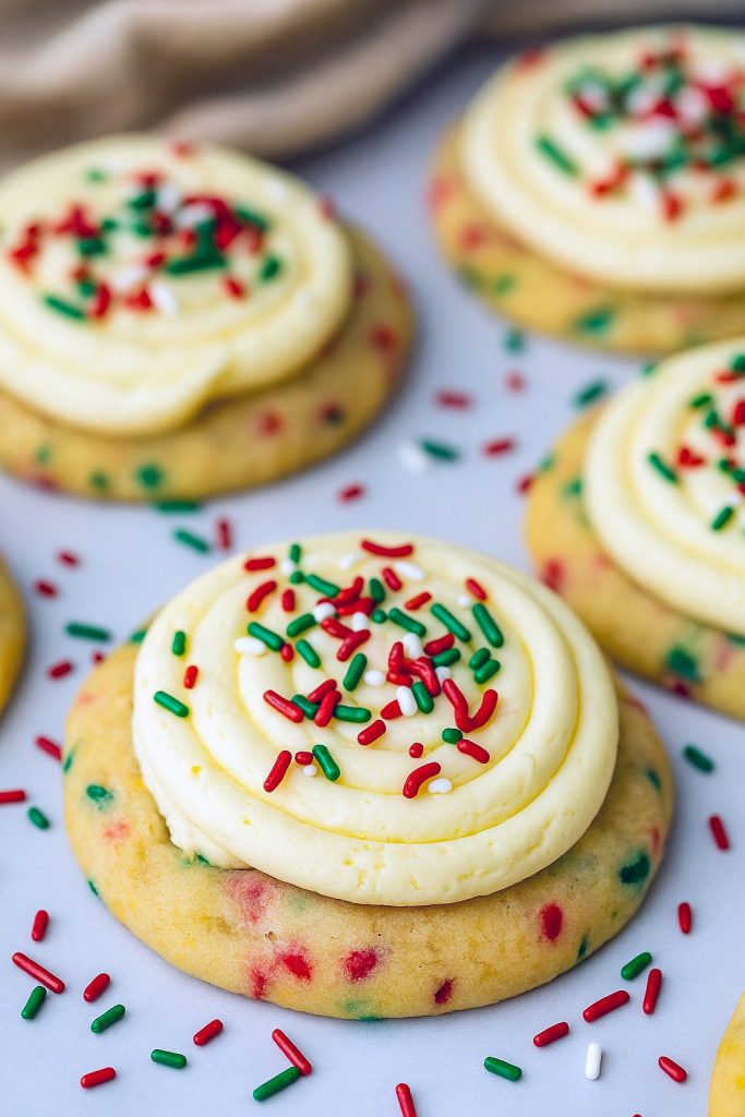 Festive stack of soft, round cake batter cookies generously coated in red, green, and white sprinkles, each topped with a swirl of buttery frosting, arranged on a holiday-themed plate.