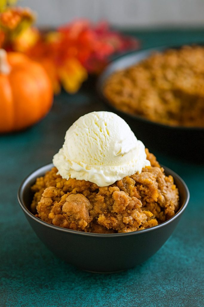 A cast-iron skillet overflowing with golden-brown Pumpkin Crisp, the perfect Pumpkin Dessert that rivals any Cobbler, set beside a stack of classic Pumpkin Dishes for a festive fall table.