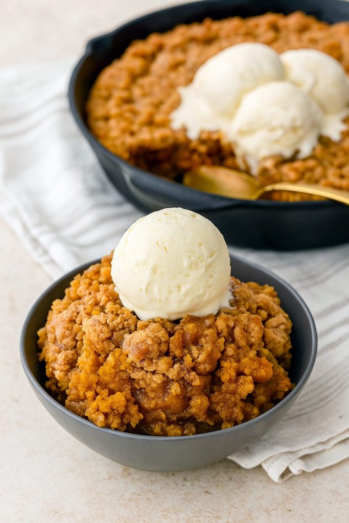 A cast-iron skillet overflowing with golden-brown Pumpkin Crisp, the perfect Pumpkin Dessert that rivals any Cobbler, set beside a stack of classic Pumpkin Dishes for a festive fall table.