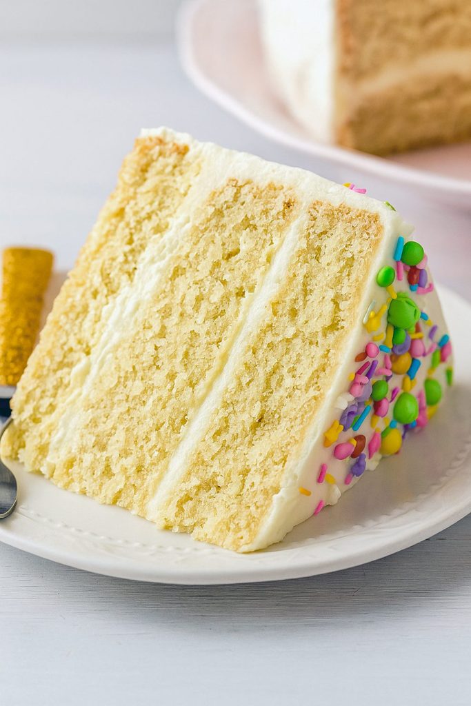 A slice of moist vanilla layer cake with smooth buttercream frosting and colorful sprinkles on a dessert plate.