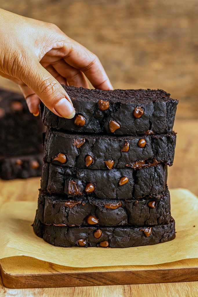 A moist, dark-brown pumpkin bread loaf studded with melted chocolate chips, sliced on a wooden cutting board with a sprig of autumn leaves nearby.