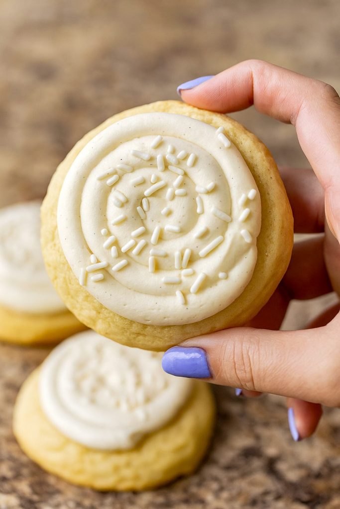Six thick vanilla-bean sugar cookies topped with swirls of creamy frosting and white sprinkles, arranged on a marble slab.