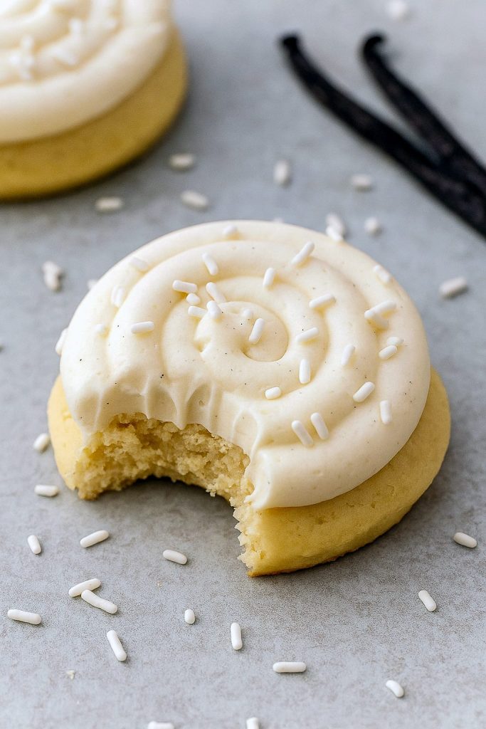 Six thick vanilla-bean sugar cookies topped with swirls of creamy frosting and white sprinkles, arranged on a marble slab.