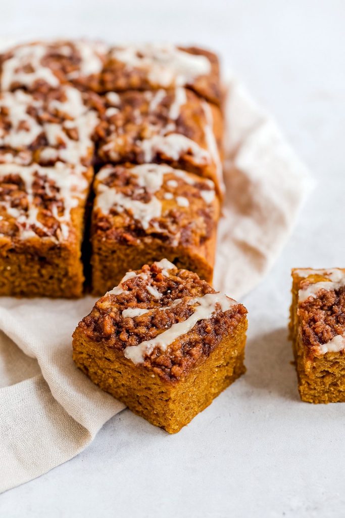 A square of pumpkin spice coffee cake topped with golden brown pecan streusel and drizzled with maple glaze, set on a rustic wooden table.