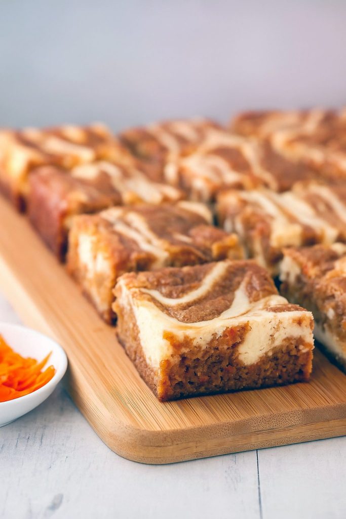 Marbled carrot cake cheesecake bars with creamy cheesecake swirls on a parchment-lined tray, topped with a dusting of cinnamon and a few toasted pecans.