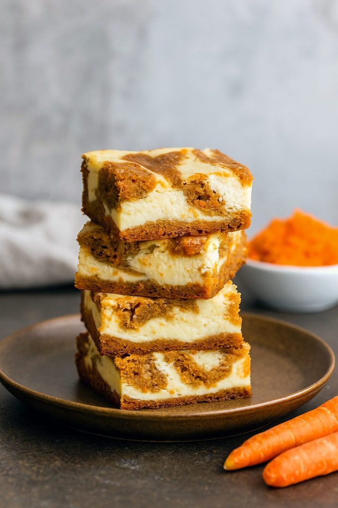 Marbled carrot cake cheesecake bars with creamy cheesecake swirls on a parchment-lined tray, topped with a dusting of cinnamon and a few toasted pecans.