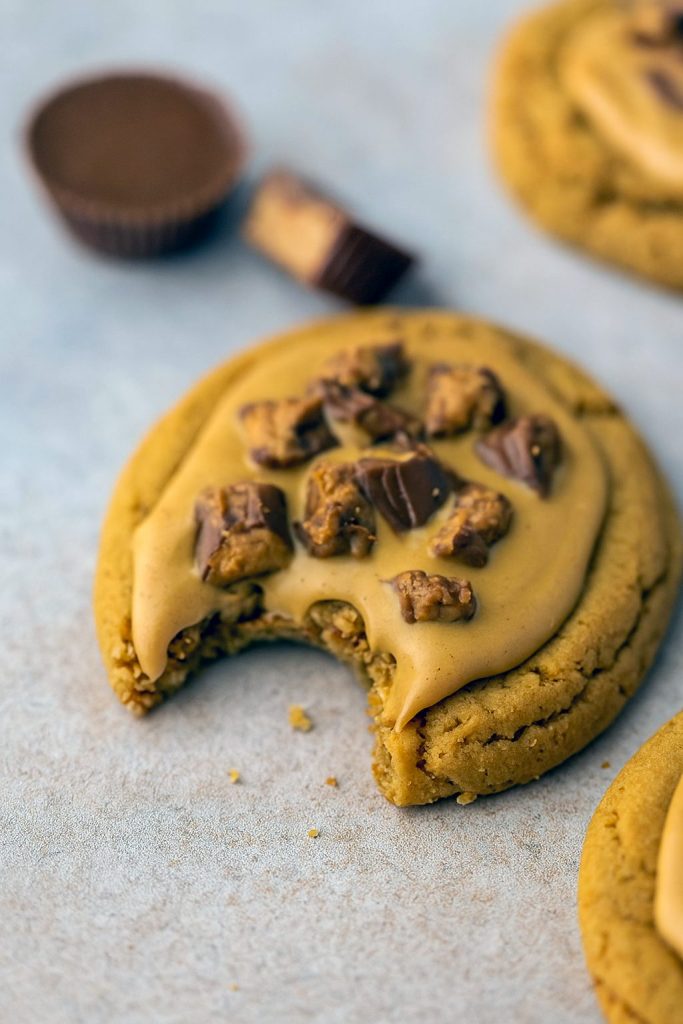 A rustic wooden board piled high with thick, chewy peanut butter cookies overflowing with chopped Reese’s peanut butter cups and drizzled with melted peanut butter chips.