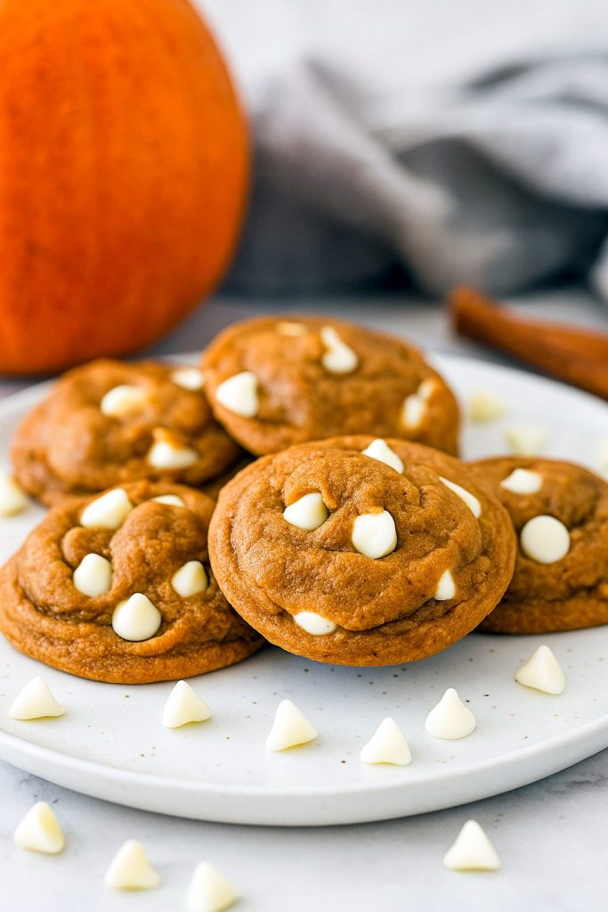 A stack of four soft, golden-brown snickerdoodle cookies speckled with white chocolate chips and pumpkin spice, topped with a dusting of cinnamon sugar.