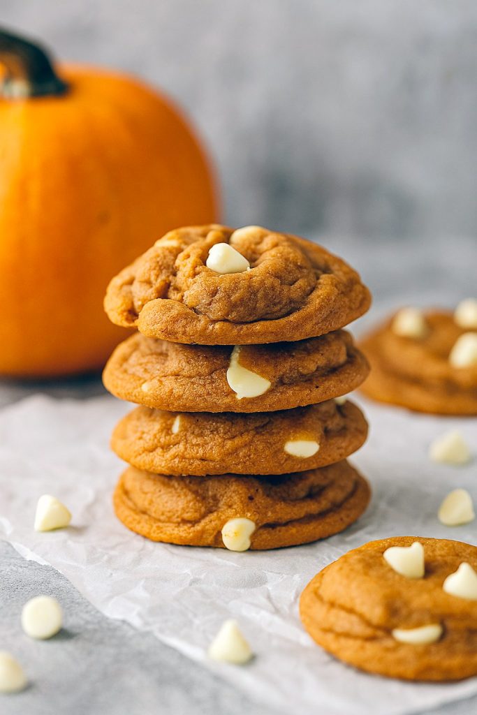 A stack of four soft, golden-brown snickerdoodle cookies speckled with white chocolate chips and pumpkin spice, topped with a dusting of cinnamon sugar.