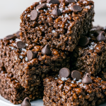 Square chocolate marshmallow cereal bars stacked on a white plate, each with melty chocolate chips and a sprinkle of sea salt, showcasing chewy and crunchy layers for a decadent treat.
