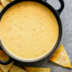 A bowl of creamy Tex Mex Queso Dip topped with cilantro and diced tomatoes, surrounded by crispy tortilla chips.