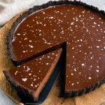 A slice of glossy dark chocolate ganache tart resting on a porcelain plate, its crisp chocolate cookie crust contrasted by a silky, mirror‑like ganache surface, dusted with flaky sea salt.