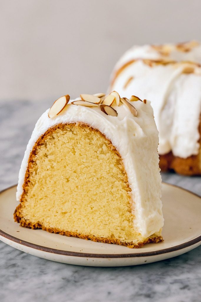 Rich and moist Amaretto Pound Cake topped with almond cream cheese frosting and slivered almonds, shown sliced on a plate with golden edges.