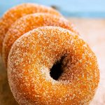 A dozen spiced apple cider–infused, cake-style donuts arranged on a cooling rack, each coated in cinnamon sugar and gleaming with autumn warmth.