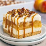 A glass dish filled with layered apple lasagna dessert: a golden graham cracker crust, creamy white cheesecake layer, juicy apple filling, topped with whipped cream and chopped pecans.