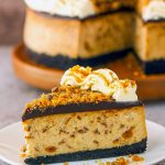 A thick slice of peanut butter-infused cheesecake in an Oreo crust, topped with glossy chocolate ganache, peanut butter whipped cream swirls, and crunchy Butterfinger pieces.