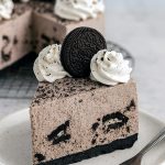 A creamy, no-bake Oreo cheesecake with a thick Oreo crust, topped with whipped cream and Oreo cookie halves, sliced to reveal the smooth filling studded with cookie bits.