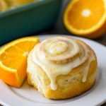 Close-up of a glossy, orange-glazed Sweet Roll spiral on a white plate, showcasing vibrant citrus zest and fluffy layers.