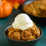 A cast-iron skillet overflowing with golden-brown Pumpkin Crisp, the perfect Pumpkin Dessert that rivals any Cobbler, set beside a stack of classic Pumpkin Dishes for a festive fall table.