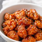 Golden sesame chicken pieces glazed in a sticky sauce, sprinkled with sesame seeds and sliced scallions, served with steamed rice and a lime wedge.