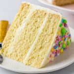 A slice of moist vanilla layer cake with smooth buttercream frosting and colorful sprinkles on a dessert plate.