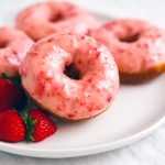 Freshly baked ring-shaped doughnuts coated in bright pink strawberry glaze, cooling on a wire rack and sprinkled with chopped strawberries.
