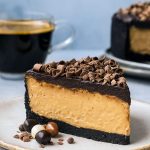A slice of creamy coffee-infused cheesecake with an Oreo crust and chocolate ganache topping, served on a white dessert plate.
