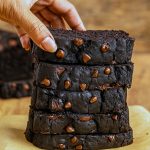 A moist, dark-brown pumpkin bread loaf studded with melted chocolate chips, sliced on a wooden cutting board with a sprig of autumn leaves nearby.
