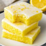 A stack of bright yellow Lemon Blondie Brownies showing moist, fudgy interiors and shiny Lemon Bars With Lemon Glaze, styled to mimic White Chocolate Lemon Brownies with drips like Lemon Brownies With White Chocolate, evoking Lemonade & Limoncello Blondies, Deserturi Raw Vegan vibes, alongside a campfire–themed Smores Dessert, surrounded by a handwritten Resep Brownies card and a layered Kek Lapis cake in the background.