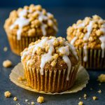Twelve golden pumpkin crumb cake muffins in a muffin tin, each topped with a crunchy buttery streusel and drizzled with maple glaze.