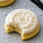 Six thick vanilla-bean sugar cookies topped with swirls of creamy frosting and white sprinkles, arranged on a marble slab.
