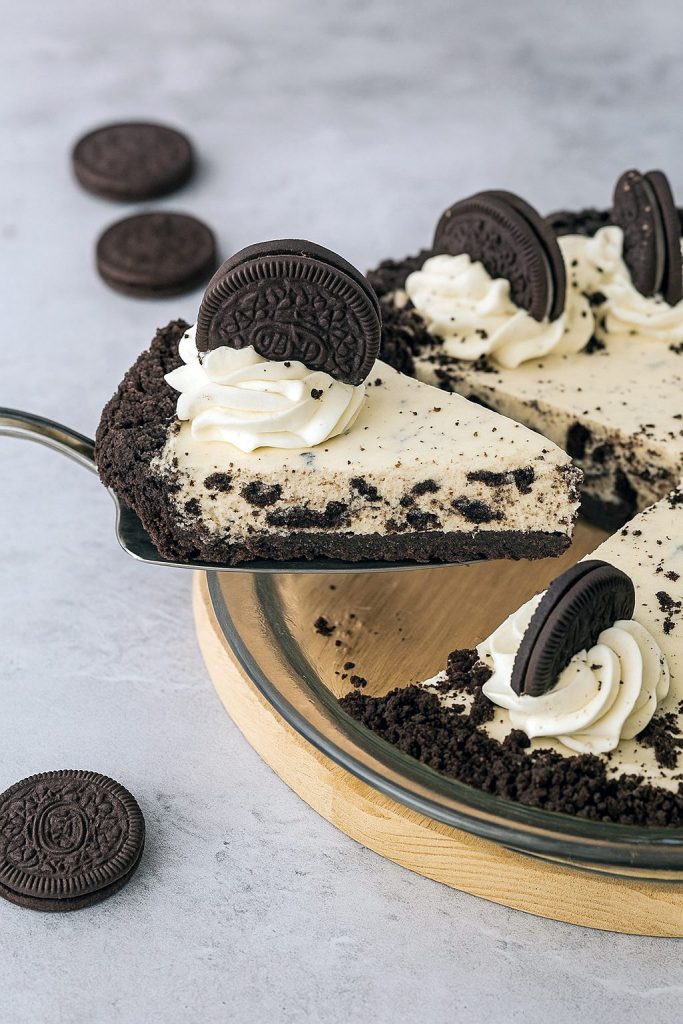 A chilled slice of no-bake Oreo pie on a white plate, showing a dark cookie crust, creamy filling flecked with chopped Oreos, and a dusting of cookie crumbs on top.