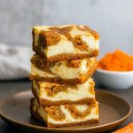 Marbled carrot cake cheesecake bars with creamy cheesecake swirls on a parchment-lined tray, topped with a dusting of cinnamon and a few toasted pecans.