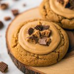 A rustic wooden board piled high with thick, chewy peanut butter cookies overflowing with chopped Reese’s peanut butter cups and drizzled with melted peanut butter chips.
