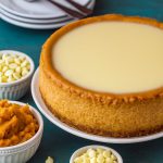 A slice of white chocolate pumpkin cheesecake with glossy white chocolate glaze and whipped cream, garnished with toasted pumpkin seeds on a rustic plate.