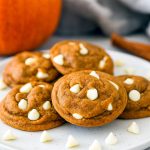 A stack of four soft, golden-brown snickerdoodle cookies speckled with white chocolate chips and pumpkin spice, topped with a dusting of cinnamon sugar.
